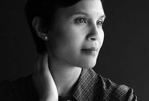 Black and white headshot of a person with short hair, looking to the side, hand touching their neck, wearing a patterned shirt.