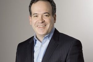 Headshot of a person wearing a dark pinstripe suit jacket and a light blue shirt, smiling against a neutral background.