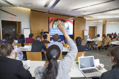 A group of people seated in a conference room, attending a presentation. One person has their hand raised, and a large screen displays a slide.