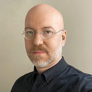 Headshot of a person with a bald head, wearing glasses, a dark shirt, and a short beard, set against a neutral background.