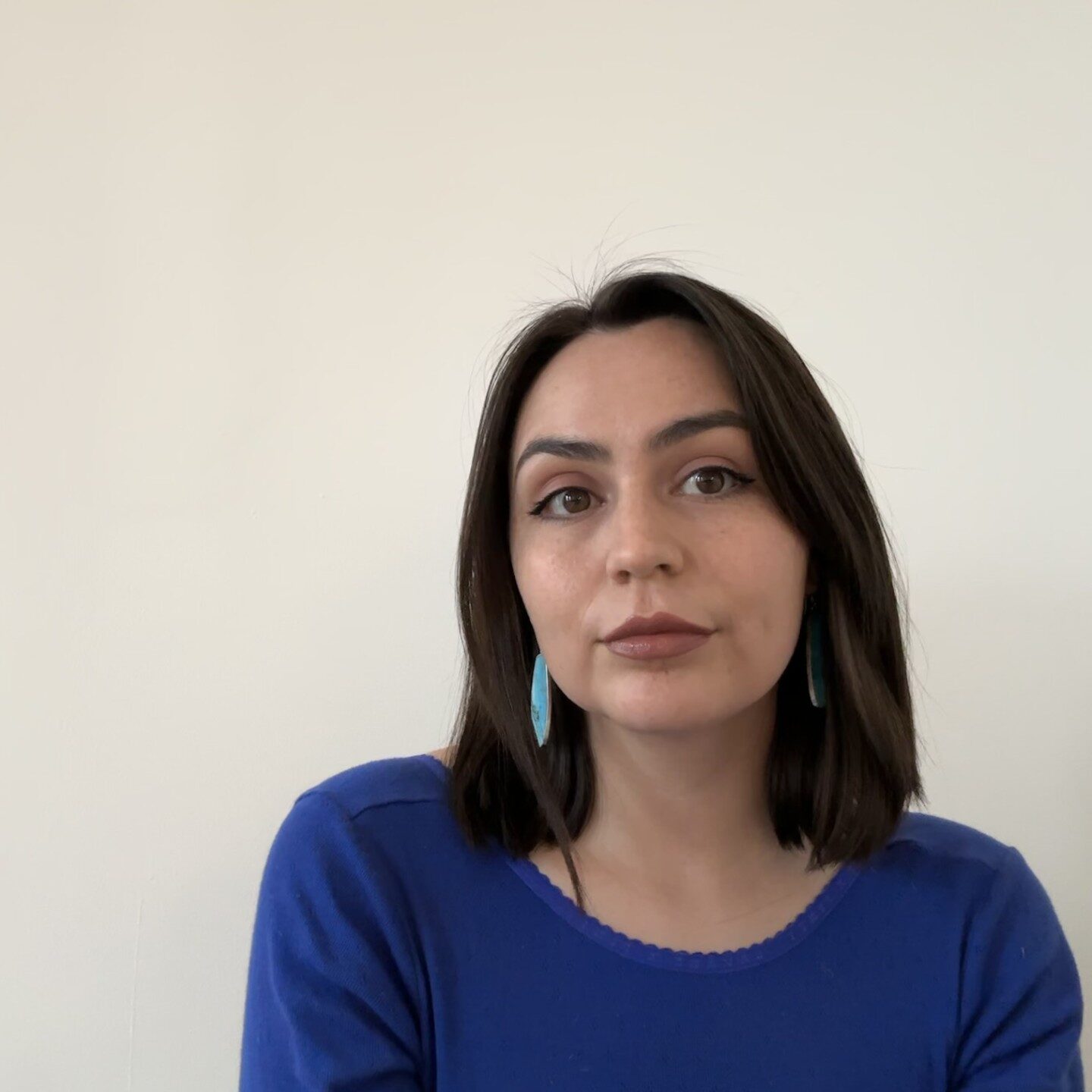 Headshot of a person with shoulder-length dark hair, wearing a blue shirt and blue earrings, set against a plain background.