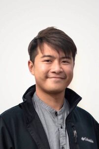 Headshot of person with short hair, wearing a dark jacket and a gray shirt, smiling at the camera.